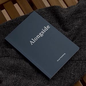 Könnte beinhalten: Ein dunkelgraues, rechteckiges Buch mit dem Wort Alongside in weißem Druck auf dem Vorderdeckel. In der unteren rechten Ecke des Einbands befindet sich der Text Wnuk Publishing in einer kleineren weißen Schrift. Das Buch hat eine matte Oberfläche.