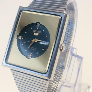Op de afbeelding: Een vintage Seiko 5 automatisch horloge met een rechthoekige goudkleurige wijzerplaat en een donkerblauwe cirkelvormige wijzerplaat. Het horloge heeft een zilverkleurige mesh band en wordt gepresenteerd op een transparante standaard. De wijzerplaat toont de dag en datum.