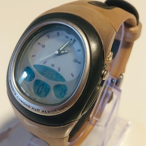 Peut inclure: Une montre de sport beige et noire avec un affichage numérique. Le cadran de la montre est clair avec des chiffres et des aiguilles noirs. Le bracelet de la montre est beige et la montre est étiquetée "SPORTS CHRONO H2O RESIST 50M".