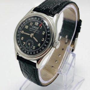 Puede incluir: Un reloj de pulsera analógico plateado con esfera negra, que incluye el texto ORIS, WATERPROOF y ANTI-SHOCK. El reloj cuenta con una aguja de fecha con punta roja, números arábigos, una subesfera y una correa de cuero negro texturizado.