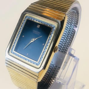 Può includere: Un orologio rettangolare color oro con quadrante blu scuro e cinturino a maglia argentata. Il quadrante dell'orologio è incorniciato da piccole pietre trasparenti. L'orologio ha il testo "ORIENT QUARTZ" sul quadrante.