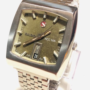 Puede incluir: Un reloj de pulsera Rado NCC 505 vintage con caja y correa plateadas. La esfera es de color verde oliva con marcadores y agujas plateadas. La fecha se muestra en la esfera. El reloj se exhibe sobre un soporte transparente.