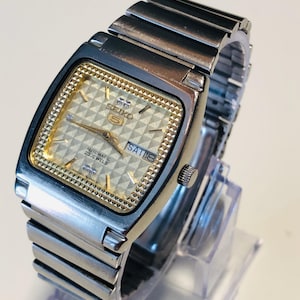 Op de afbeelding: Een zilverkleurig Seiko 5 automatisch polshorloge met een vierkante kast en een gestructureerde goudkleurige wijzerplaat met een geometrisch patroon. Het horloge heeft een dag-datumvenster op de drie-uurpositie, gouden wijzers en een metalen schakelarmband.