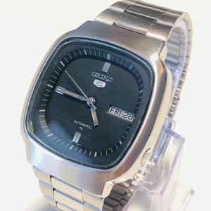May include: A Seiko 5 automatic wristwatch with a square-shaped silver-tone stainless steel case and matching link bracelet. The watch features a black dial with silver-tone hour markers, silver hands, and a day-date window displaying FRI 28.