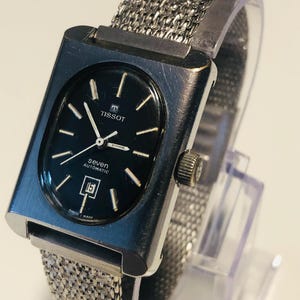 May include: A vintage Tissot Seven automatic watch with a rectangular silver case and a black dial. The watch has silver hands and markers, a date window, and a mesh metal strap. The watch is Swiss made.