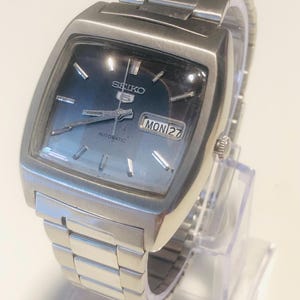Peut inclure: Une montre-bracelet automatique Seiko 5 argentée avec un boîtier carré et un bracelet à maillons en acier inoxydable assorti. La montre présente un cadran bleu dégradé, des index argentés et un guichet jour-date affichant MON 27.