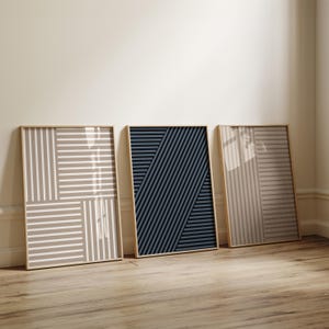 May include: Three framed abstract art prints. The prints feature geometric designs with horizontal and diagonal lines in neutral tones of beige, gray, and navy blue. The frames are a light wood color, and the prints are leaning against a wall.