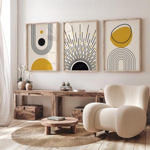 May include: Three framed abstract art prints with geometric designs in black, yellow, and beige. The prints feature sunburst, arch, and circle shapes. The artwork is displayed above a wooden console table in a modern living room setting.
