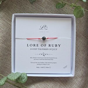 May include: A red string bracelet featuring a central faceted dark stone bead flanked by two small gold-coloured beads, presented on a white card labelled LORE OF RUBY LUCKY TALISMAN OF JULY with printed instructions and the Loom & Lore logo.