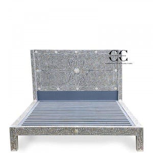 Op de afbeelding: Een handgemaakt bedframe met een decoratief hoofdbord. Het hoofdbord en frame zijn versierd met ingewikkelde bloemen- en bladerpatronen in een lichte kleur, met een blauw accent. Het bedframe heeft een lattenbodem.