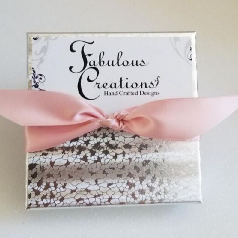 May include: A white gift box with a silver and white patterned design. The box is tied with a pink satin ribbon and has a label that reads "Fabulous Creations Hand Crafted Designs".