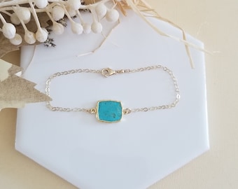 May include: A delicate gold-toned bracelet featuring a square, turquoise-colored stone at its center. The stone is set in a gold bezel and is connected to a fine chain. The bracelet is displayed on a white, hexagonal surface.