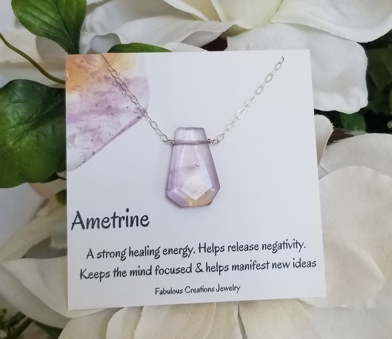 May include: Ametrine necklace with a purple and orange crystal pendant. The necklace is on a white card with the text "Ametrine", "A strong healing energy. Helps release negativity. Keeps the mind focused & helps manifest new ideas", and "Fabulous Creations Jewelry".