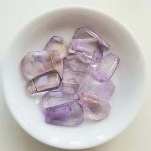 May include: A collection of eight faceted amethyst gemstone beads in a white bowl. The beads are various shades of purple and have a translucent appearance.