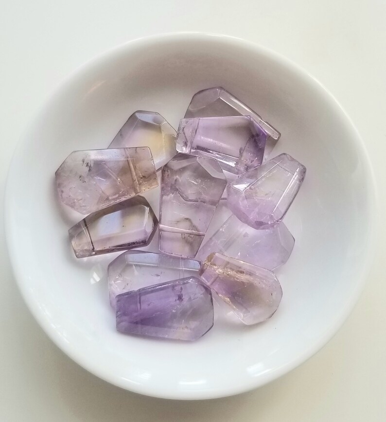 May include: A collection of eight faceted amethyst gemstone beads in a white bowl. The beads are various shades of purple and have a translucent appearance.