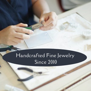 May include: A close-up of a person's hands working on a piece of jewelry. The person is using pliers to attach a small, clear crystal to a silver chain. The image is set against a white background with tools and beads scattered around. The text "Handcrafted Fine Jewelry Since 2010" is displayed in a black oval.