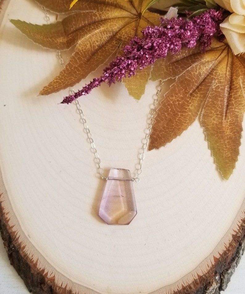 May include: A delicate silver chain necklace with a faceted, teardrop-shaped amethyst pendant. The pendant is a light purple color with hints of pink and yellow.