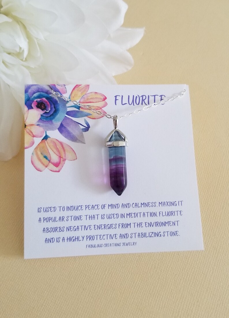 May include: A silver chain necklace with a purple and blue fluorite crystal pendant. The pendant is shaped like a point and has a silver cap. The necklace comes with a card that reads "Fluorite is used to induce peace of mind and calmness. Making it a popular stone that is used in meditation. Fluorite absorbs negative energies from the environment and is a highly protective and stabilizing stone. Fabulous Creations Jewelry."