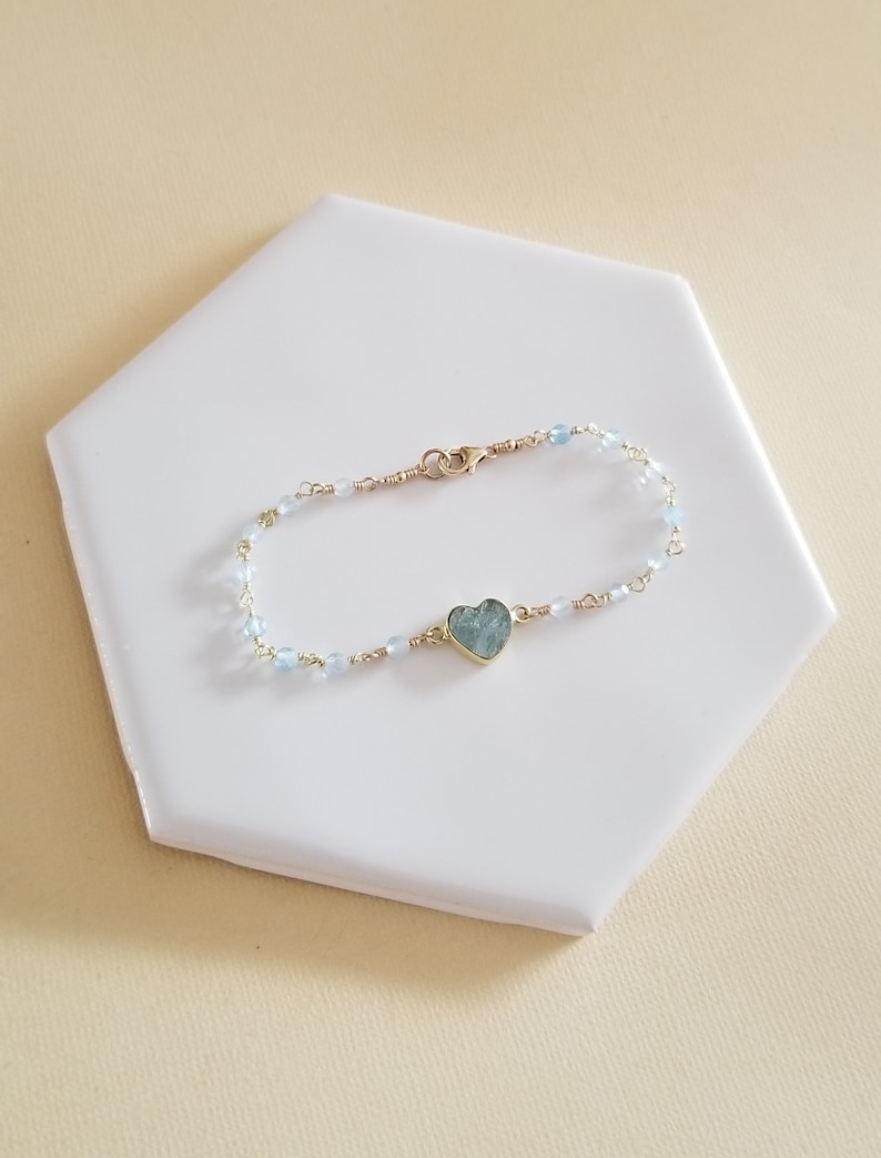 May include: A delicate gold chain bracelet with light blue beads and a heart-shaped aquamarine pendant.