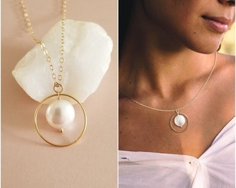 May include: A gold chain necklace with a delicate circle pendant featuring a white pearl. The necklace is displayed on a white rock and on a model wearing a white shirt.