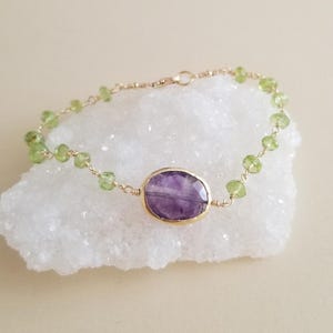 May include: Gold bracelet featuring a large, oval amethyst gemstone in the center, surrounded by smaller, round peridot stones. The bracelet is set on a white surface, showcasing the contrast of colors and textures.