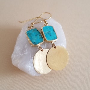 May include: A pair of gold-toned drop earrings featuring a rectangular turquoise stone set in a gold-coloured frame, attached to a circular, hammered gold-toned metal disc. The earrings include standard gold-toned wire hooks for attachment.