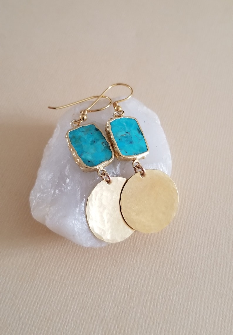 May include: A pair of gold-toned drop earrings featuring a rectangular turquoise stone set in a gold-coloured frame, attached to a circular, hammered gold-toned metal disc. The earrings include standard gold-toned wire hooks for attachment.