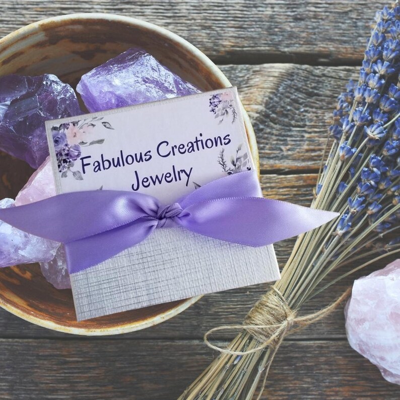 May include: A small, white gift box with a purple ribbon tied in a bow. The box has the text "Fabulous Creations Jewelry" printed on it. The box is sitting on a wooden surface with purple and pink crystals and a bundle of dried lavender.