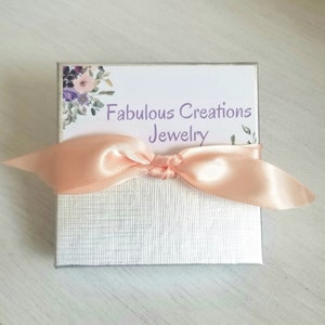 May include: A silver gift box with a peach-colored satin ribbon tied around it. The box has a floral design on the top and the text "Fabulous Creations Jewelry" printed on a white label.