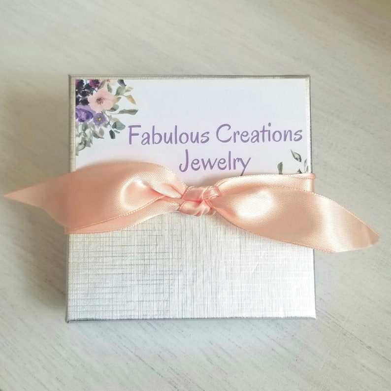 May include: A silver gift box with a peach-colored satin ribbon tied around it. The box has a floral design on the top and the text "Fabulous Creations Jewelry" printed on a white label.