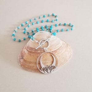 May include: A silver necklace with a turquoise bead chain and a circular pendant featuring a crescent moon and floral design. The necklace is displayed on a seashell.
