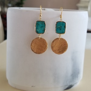 May include: A pair of drop earrings featuring a rectangular turquoise stone set in gold-toned metal above a circular, hammered gold-toned metal disc. The earrings hang from gold-toned ear wires, showcasing a contrast between the blue stone and the textured gold finish of the circular components.