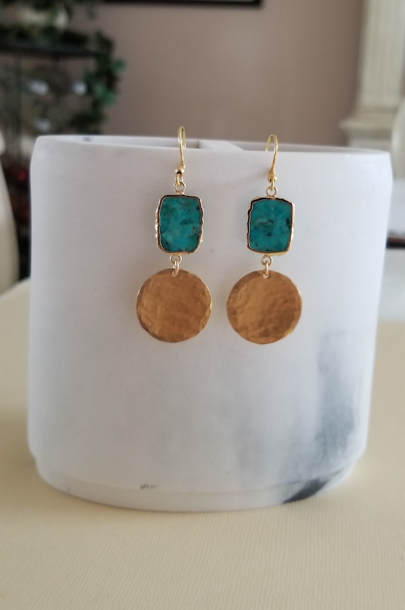 May include: A pair of drop earrings featuring a rectangular turquoise stone set in gold-toned metal above a circular, hammered gold-toned metal disc. The earrings hang from gold-toned ear wires, showcasing a contrast between the blue stone and the textured gold finish of the circular components.