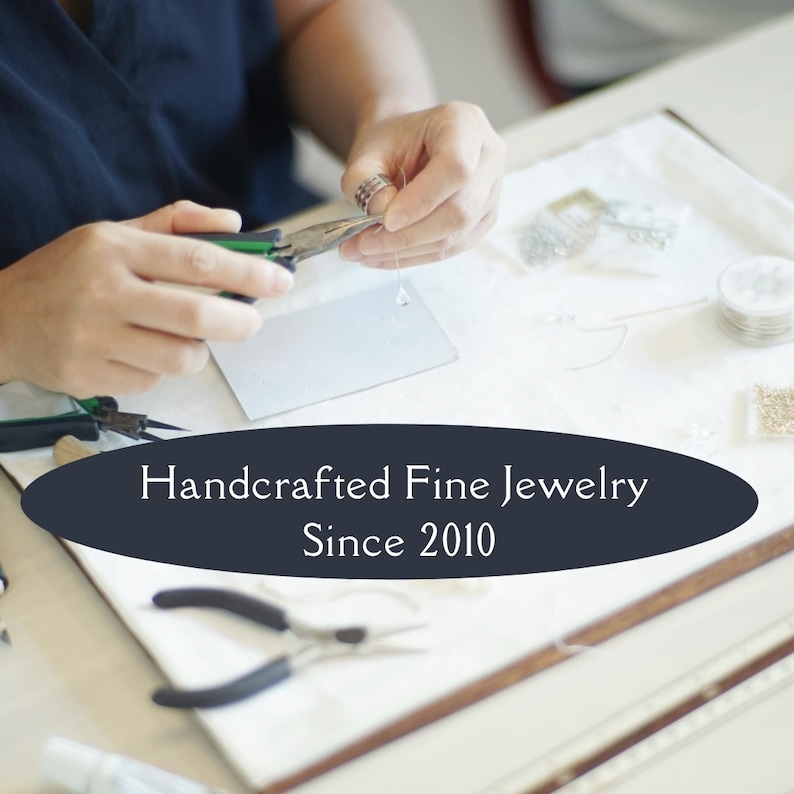 May include: A pair of metal jewellery pliers with green handles being used to assemble a delicate earring with a clear crystal bead. The image features a dark oval overlay with the white text Handcrafted Fine Jewelry Since 2010 centred on the display.