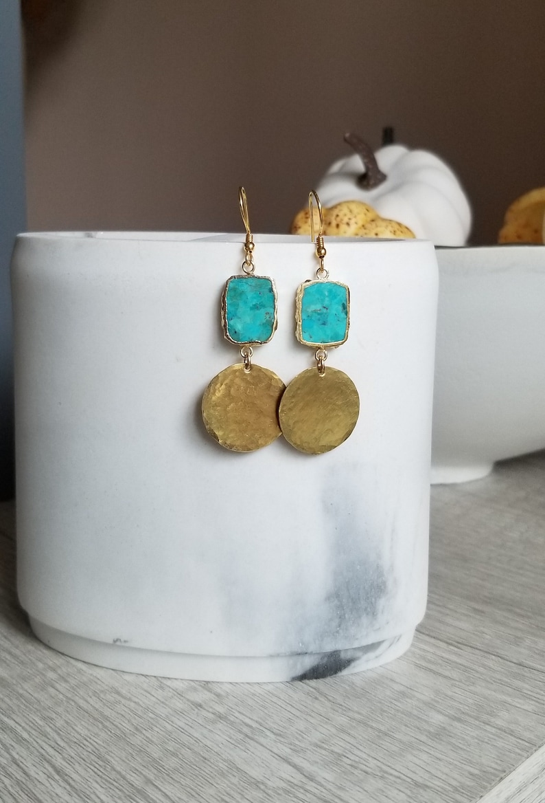May include: Dangle earrings featuring a rectangular turquoise stone set in gold-toned metal above a circular, hammered gold-toned metal disc. The earrings hang from gold-toned wire hooks, showcasing a contrast between the bright blue stone and the textured metallic finish of the circular drops.