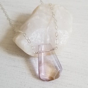 May include: A silver chain necklace with a faceted, teardrop-shaped amethyst pendant. The pendant is a light purple color with a hint of yellow.