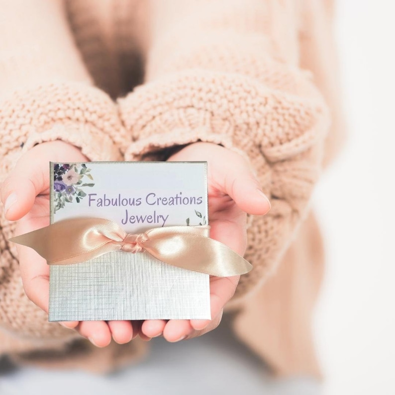 May include: A square silver jewellery box featuring a white label with the text Fabulous Creations Jewelry and a floral illustration in the corner. The box is tied with a smooth, light pink satin ribbon across the centre of the lid.