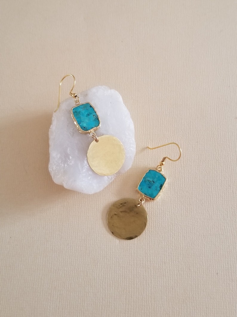 May include: A pair of drop earrings featuring a rectangular turquoise stone set in a gold-toned metal frame, attached to a circular, hammered gold-toned metal disc. The earrings hang from gold-toned metal ear wires.