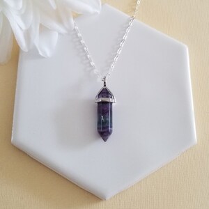 May include: A silver chain necklace with a purple and green fluorite crystal pendant. The pendant is shaped like a point and has a silver setting.
