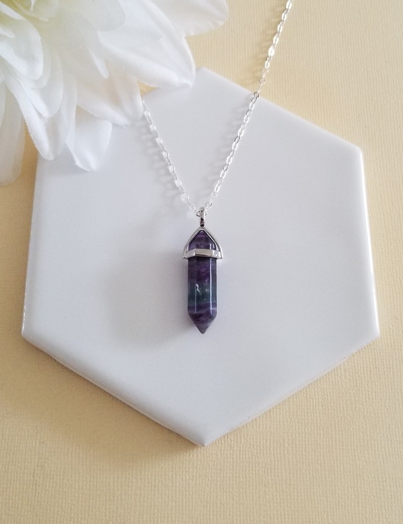 May include: A silver chain necklace with a purple and green fluorite crystal pendant. The pendant is shaped like a point and has a silver setting.