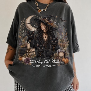 May include: A charcoal grey t-shirt featuring a graphic print of a person in a witch hat surrounded by three black cats, a crescent moon, books, pumpkins, and a potion bottle. The text Witchy Cat Club is printed at the bottom in white script.