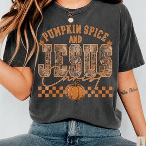 May include: A charcoal gray short-sleeve t-shirt featuring the orange text PUMPKIN SPICE AND JESUS CHRIST. The word JESUS is filled with a camouflage pattern. Below the text is an orange pumpkin graphic and an orange and gray checkerboard pattern.