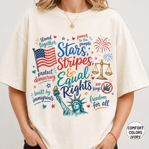 May include: Ivory-coloured t-shirt with a patriotic design featuring the American flag, Statue of Liberty, and text that reads "Stars, Stripes, & Equal Rights." The design includes fireworks, scales, and other patriotic symbols.