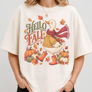 May include: An off-white t-shirt featuring a graphic of a white duck wearing a yellow cable-knit sweater, a red scarf, and orange rain boots. The text "Hello Fall" is printed in green and orange, surrounded by orange pumpkins and autumn leaves.
