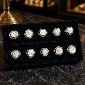 May include: Ten silver rings with white spiral centres are displayed in a black velvet box. The rings have different silver band designs, including beaded and smooth styles. The rings are presented in a black velvet jewellery box.
