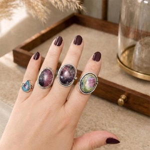 May include: A hand shows four rings with gemstone settings. The rings have oval and teardrop-shaped stones in pink, purple, and blue, set in silver bands. The gemstones have unique patterns and textures, showcasing natural variations.