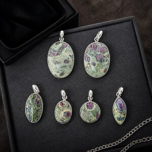 May include: Six gemstone pendants with silver bails are displayed in a black jewellery box. The pendants feature oval, round, and teardrop shapes with green and red-purple colours. A silver chain is visible.