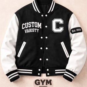 Puede incluir: Chaqueta universitaria negra con mangas blancas. La chaqueta presenta el texto "CUSTOM VARISTY" y una gran "C" blanca en el pecho. Rayas blancas adornan los puños. La manga muestra "Est. 2022".
