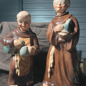 May include: Two ceramic monk figurines. The figures wear brown robes with tan rope belts and hold blue-green pitchers and cups. The monks have bald heads and smiling faces. The figurines are on a dark brown surface.
