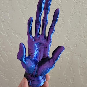 May include: A 3D-printed sculpture of a hand, with a metallic purple and blue color gradient. The hand is open, with long, slender fingers. The sculpture is mounted on a circular base with a detailed design.